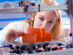 Das Bild zeigt eine Frau, die einen 3D-Drucker bei der Arbeit beobachtet.