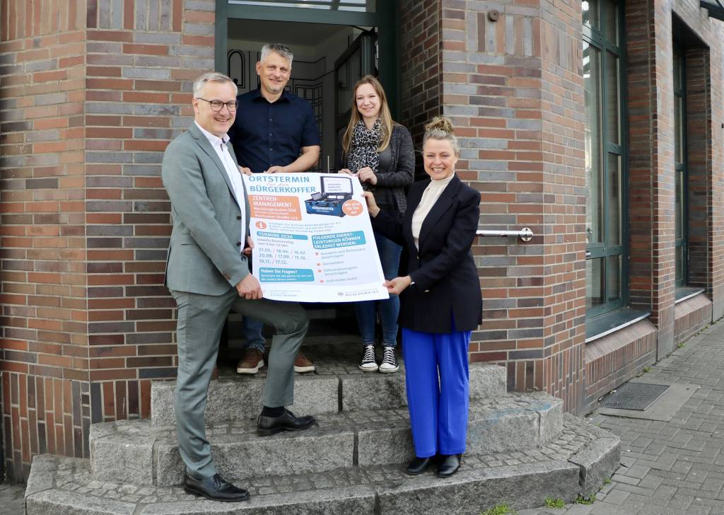 B&uuml;rgermeister Axel Tschersich pr&auml;sentiert gemeinsam mit der Ersten Beigeordneten Silke Ehrbach-Wulfen (r.) und Tim Berens (2.v.l.) sowie Corina Lehrich vom B&uuml;rgerb&uuml;ro das Plakat zum neuen B&uuml;rgerkoffer-Angebot, das in der Bochumer Stra&szlig;e 123 (ehemals Commerzbank) beheimatet ist. Foto: Stadt RE  